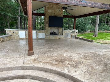 Covered outdoor patio with a stone fireplace, mounted TV, built-in kitchen cabinets, and stamped concrete flooring.