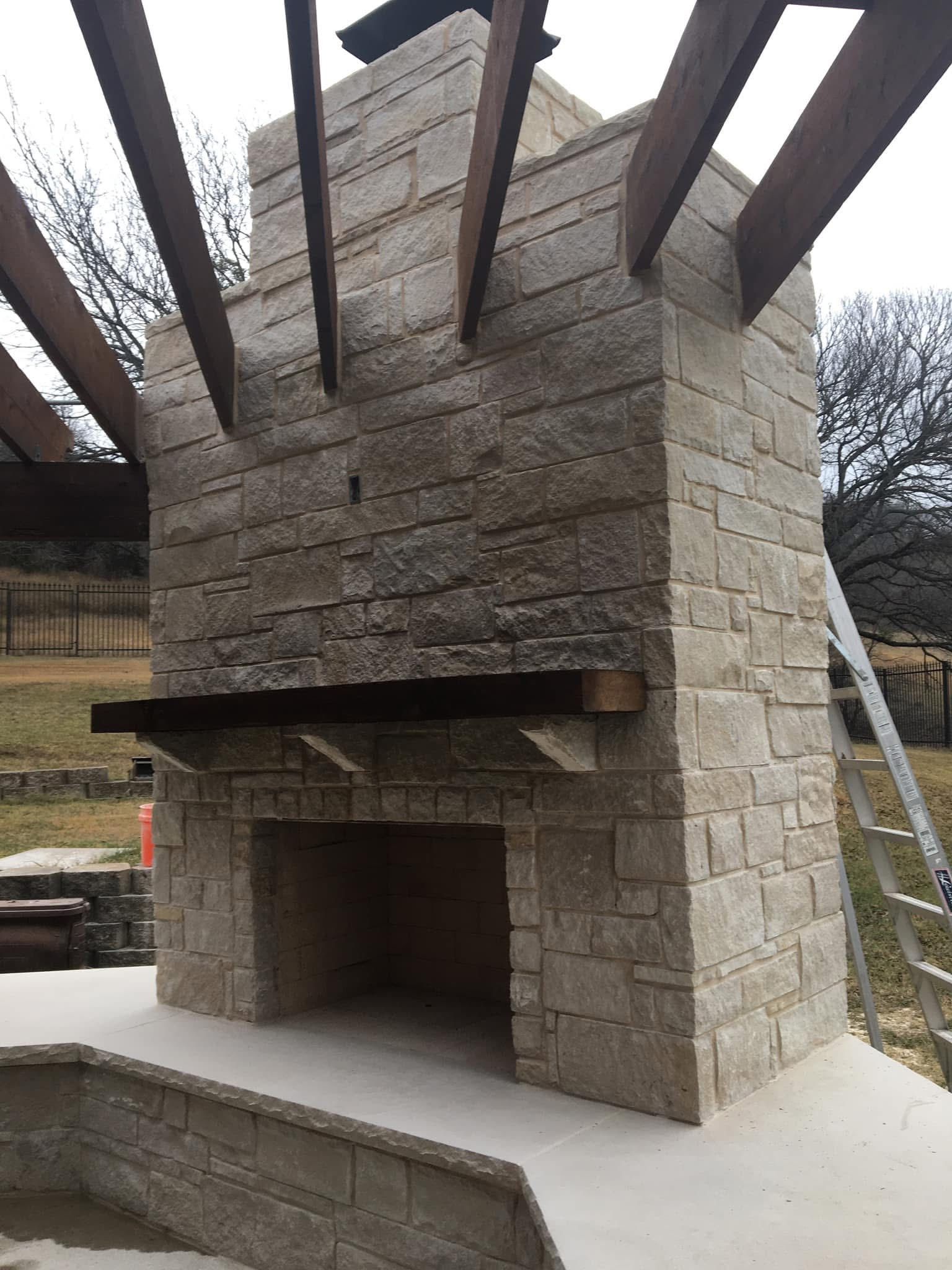 A large, light-colored stone outdoor fireplace with a wooden mantel and timber pergola frame in a backyard setting.