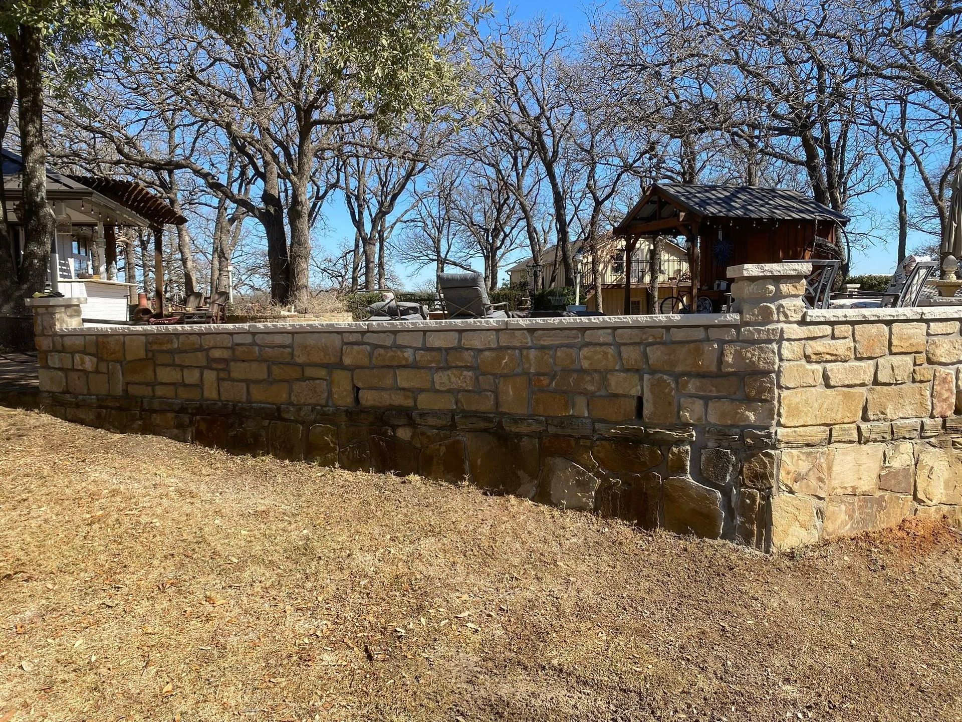 A tan stone retaining wall borders a patio area, with bare trees and structures visible in the background under blue sky.