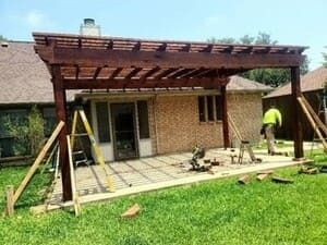 A worker builds a large, dark wood pergola over a brick patio behind a residential home.