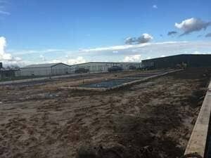 A construction site with cleared, muddy ground, a concrete foundation in the center, and metal buildings in the background.