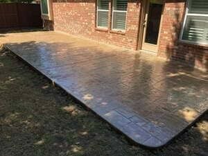 A newly poured, stamped concrete patio in a tan pattern sits adjacent to a red brick house wall.