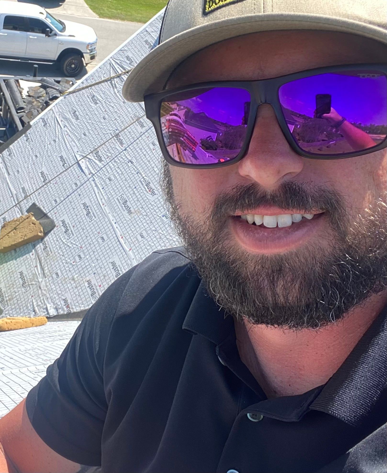 A smiling person with a beard wearing a baseball cap and purple-tinted sunglasses, outdoors on a slanted rooftop.