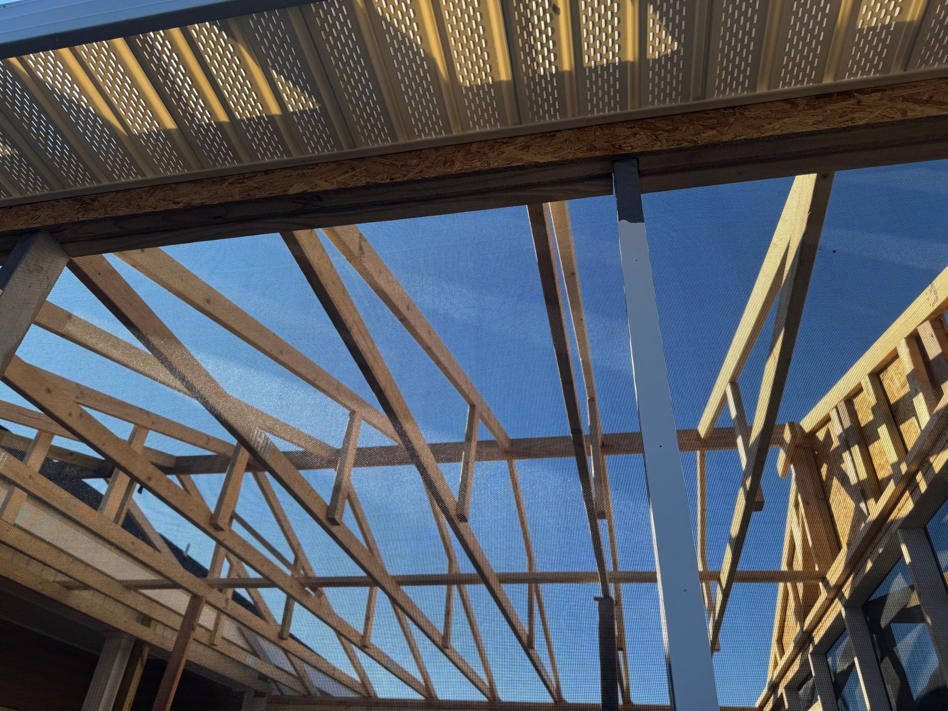 A view looking up at unfinished wooden roof trusses and an exterior soffit against a blue sky.