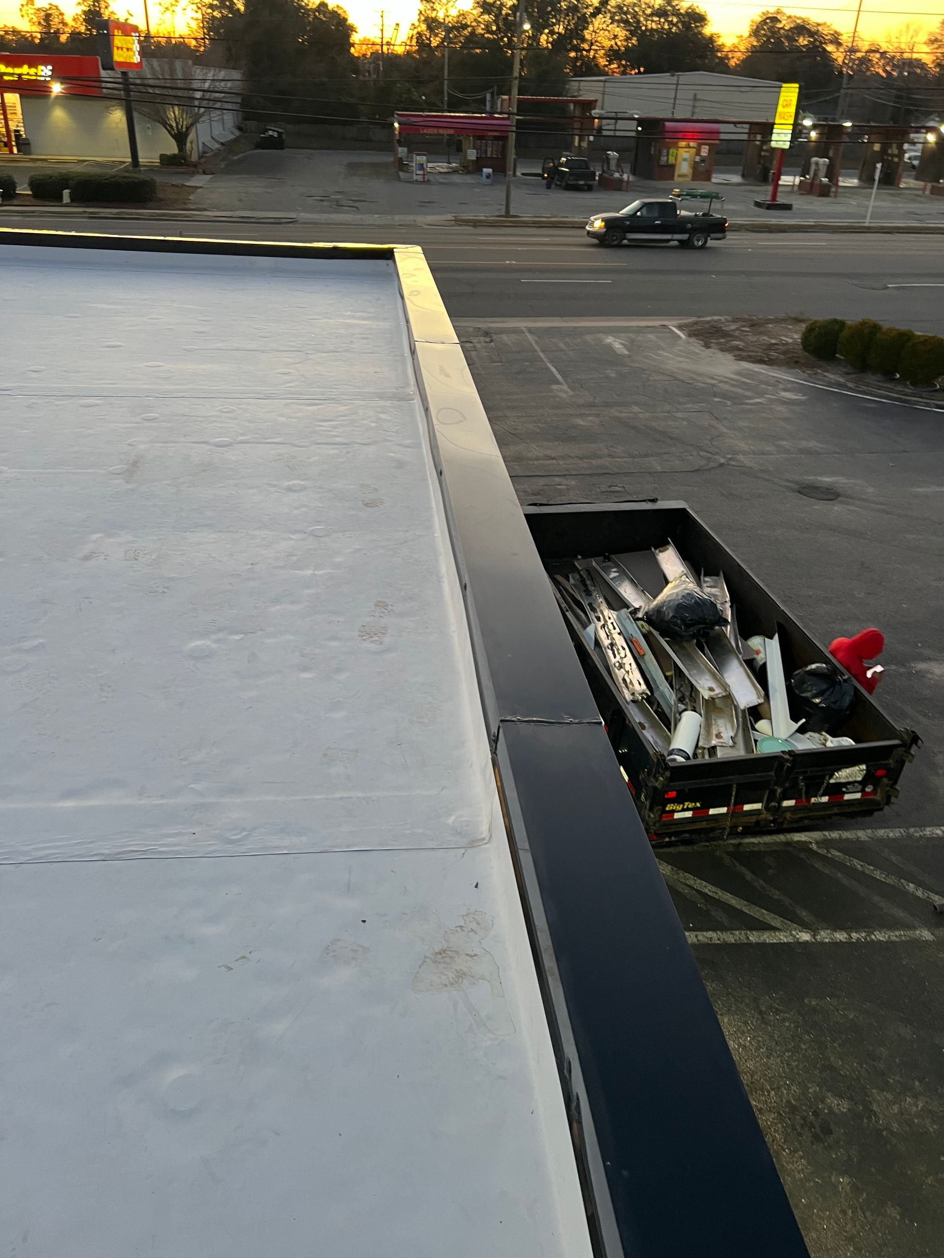A view from a flat, light-colored commercial rooftop looking down at a metal dumpster filled with construction debris.