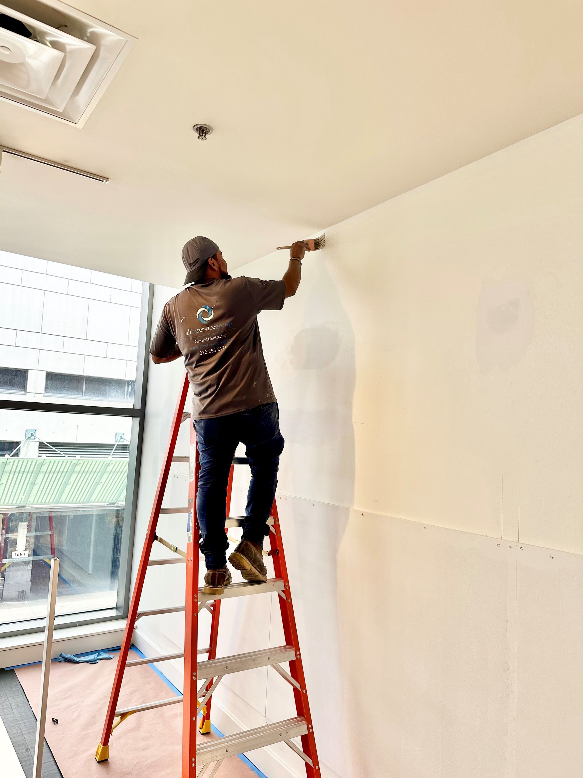 Person on a red ladder painting a white wall indoors near a window.