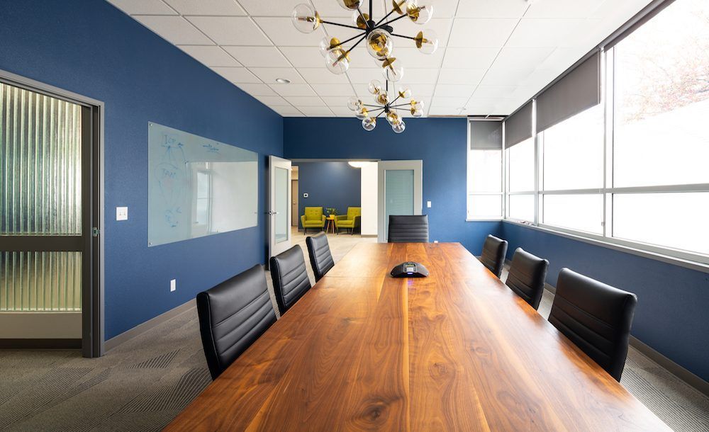 Modern conference room with a long wooden table, black chairs, blue walls, and large windows.