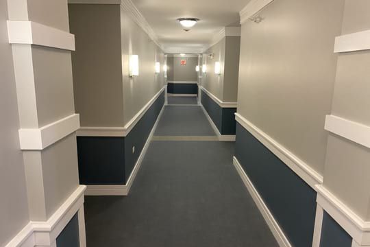 Long, empty hallway with beige walls, dark blue wainscoting, and carpeted floor under ceiling lights