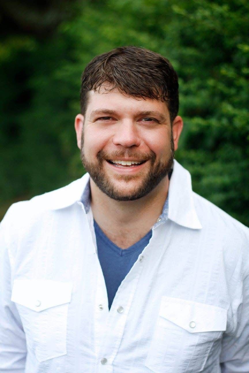 A man with a beard is wearing a white shirt and smiling.