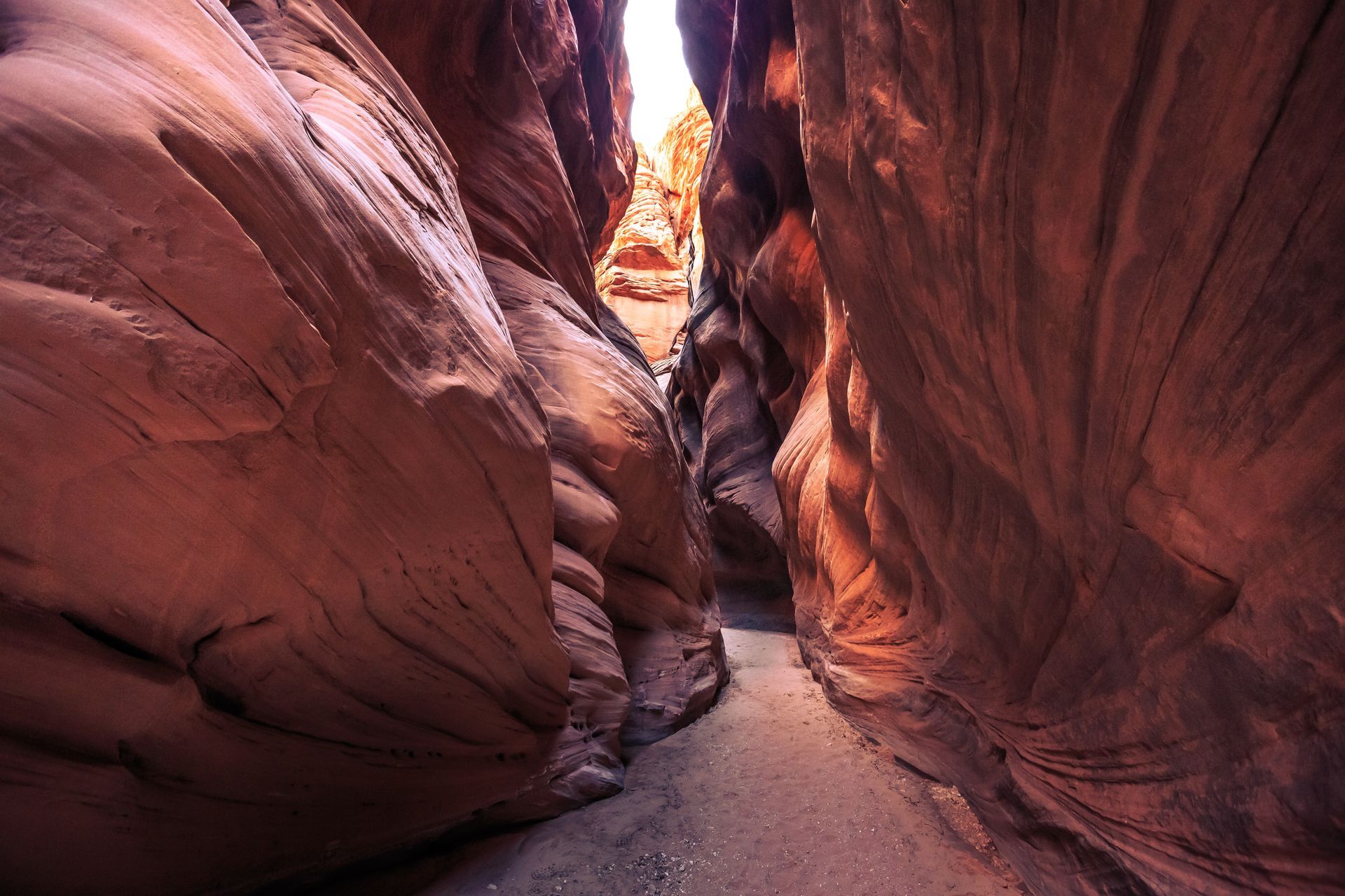 Narrow sandstone canyon with red rock walls and a sandy path lit by warm sunlight