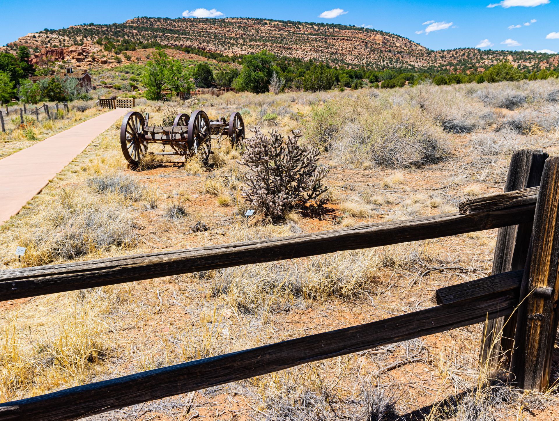 Desert trail with wooden fence, old wagon wheels, and red rock hills under a bright sky