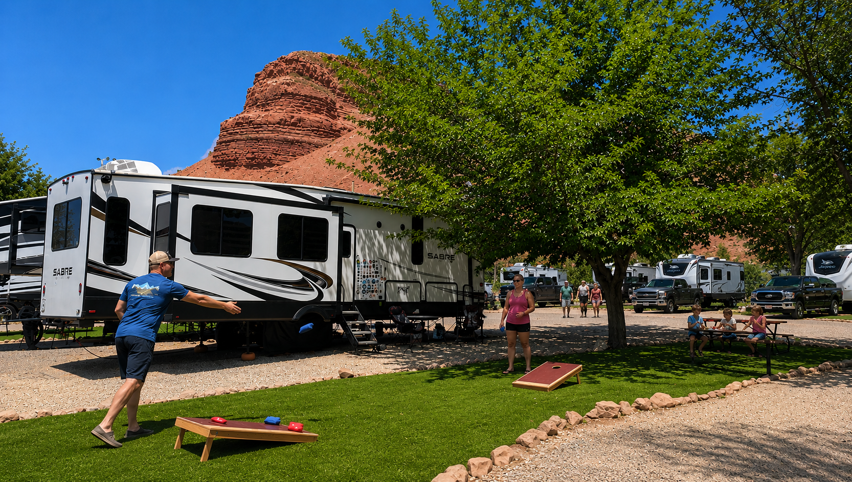 White travel trailer at a gravel campsite beneath trees, with red cliffs and blue sky in the background
