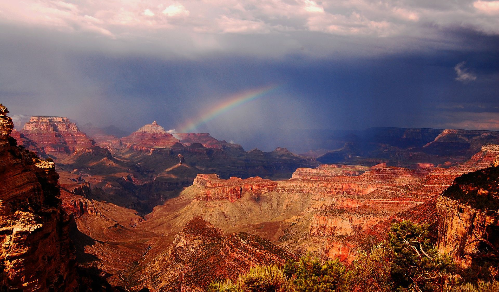 Grand Canyon at sunset with dramatic clouds and a faint rainbow over red-rock cliffs