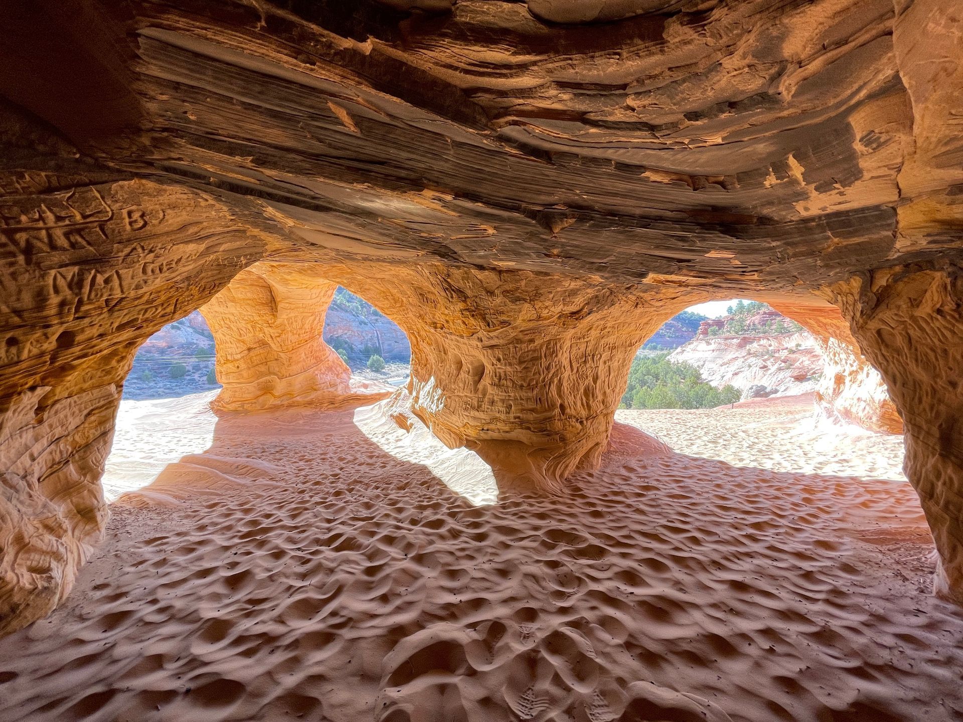 Sandstone cave opening onto a sunlit beach with rippled sand and blue sky visible outside