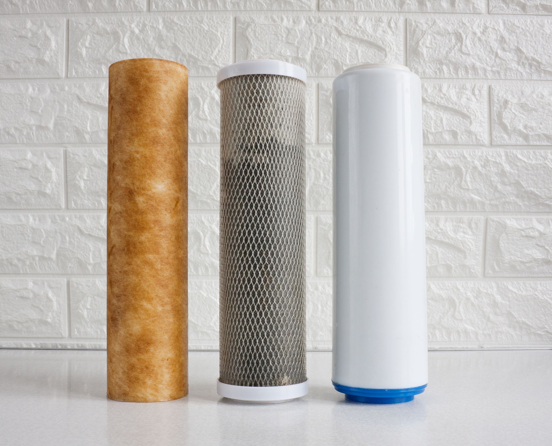 Three cylindrical water filters: tan, gray mesh, and white with blue base on a white brick background
