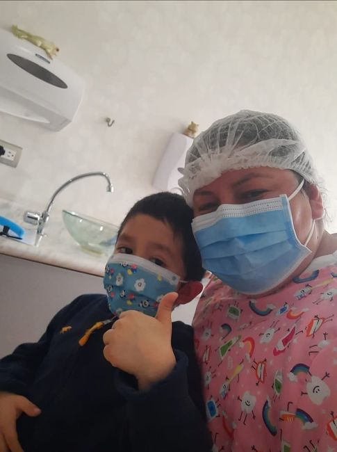 Dos personas con mascarillas posan juntas en una sala de clínica con un lavabo y suministros médicos.