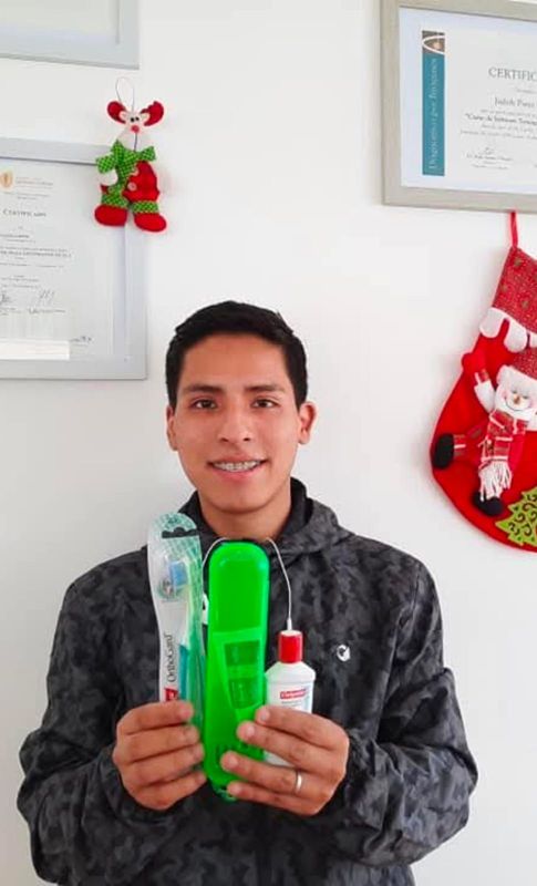 Un hombre sostiene un objeto verde y una botella con tapón rojo en un espacio cerrado, con decoraciones festivas al fondo