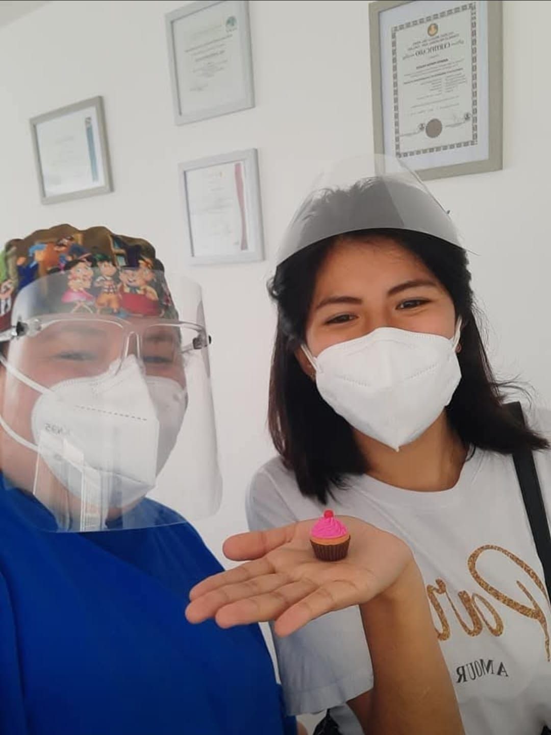 Dos personas con mascarillas y pantallas faciales en una clínica, sosteniendo un pequeño dulce rosa en la palma de la mano