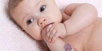 Bambina sdraiata su una coperta rosa, che guarda la macchina fotografica con le dita in bocca.