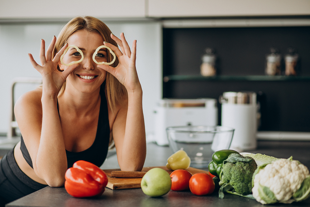 Donna sorridente in cucina, con in mano fette di cetriolo a forma di occhi e verdure fresche sul bancone.