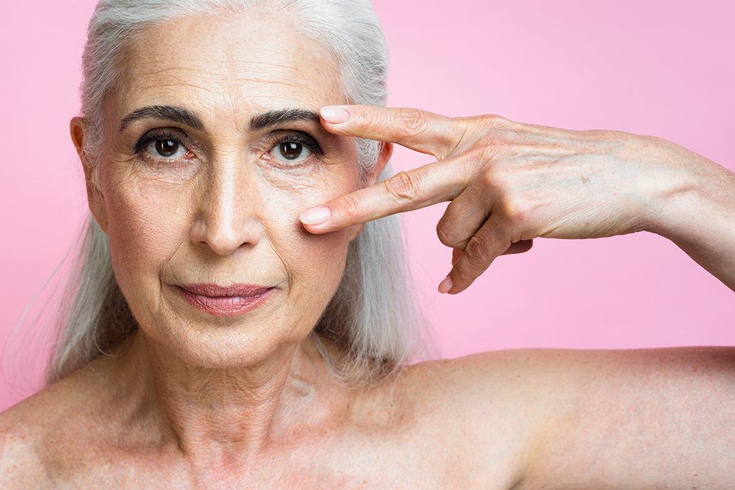 Persona con i capelli argentati in posa su uno sfondo rosa, con due dita vicino a un occhio