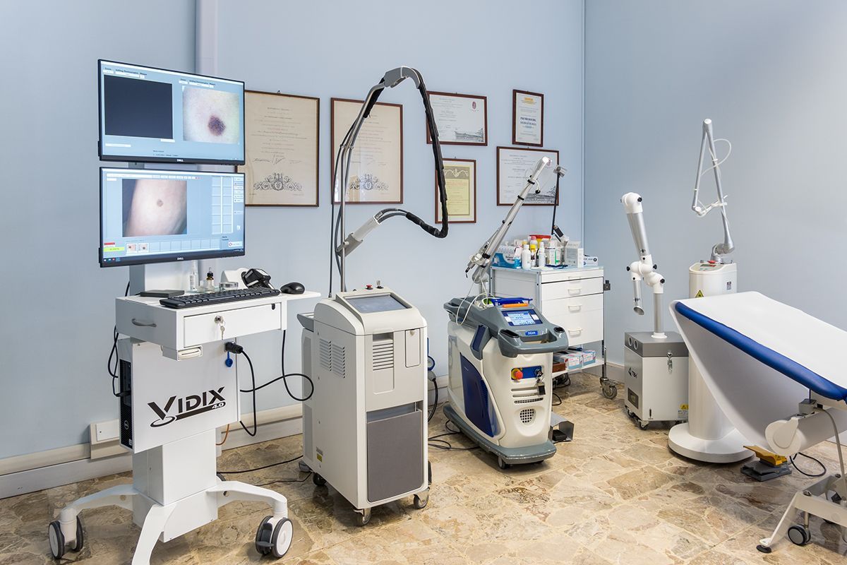 Sala visite mediche con monitor, carrelli per apparecchiature e poltrona per trattamenti, in un ambiente clinico pulito.