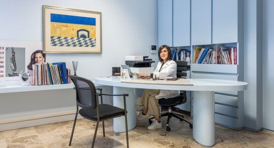Ufficio moderno con due persone sedute a delle scrivanie, una delle quali a una grande scrivania bianca, e librerie sullo sfondo.