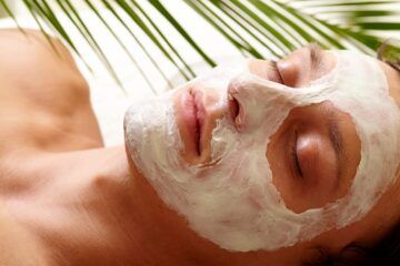 Persona che riceve una maschera facciale bianca durante un trattamento in una spa, con gli occhi chiusi e foglie di palma sopra la testa.