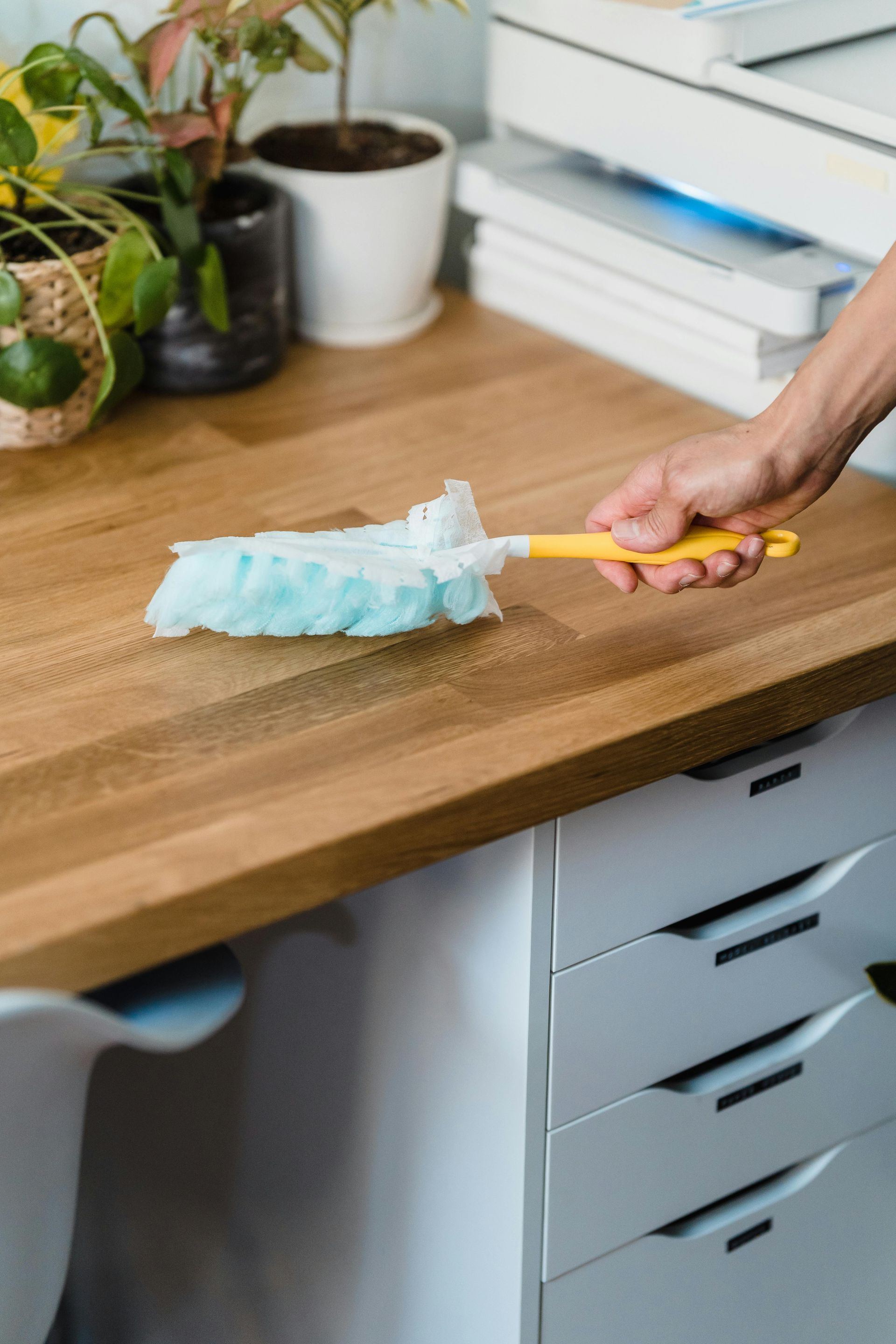 Person dusting a wooden desk with a light blue and yellow duster; white drawers and plants in the background.