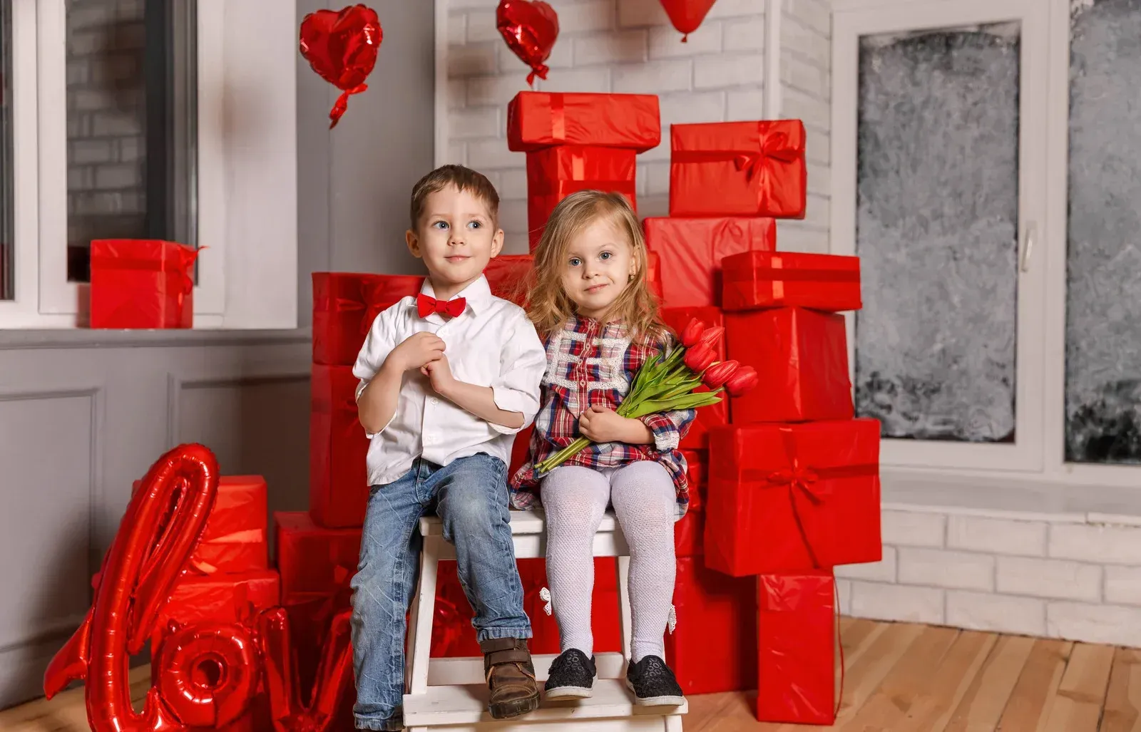 A boy and a girl are sitting on a chair in front of a pile of red gifts.