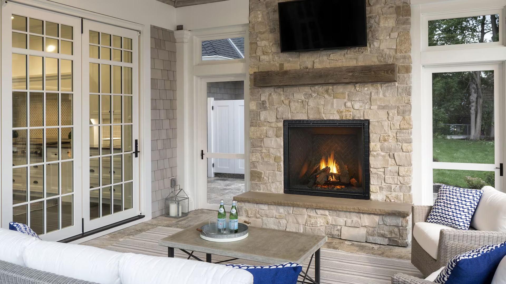 Outdoor living space with stone fireplace, TV, seating, and glass doors.