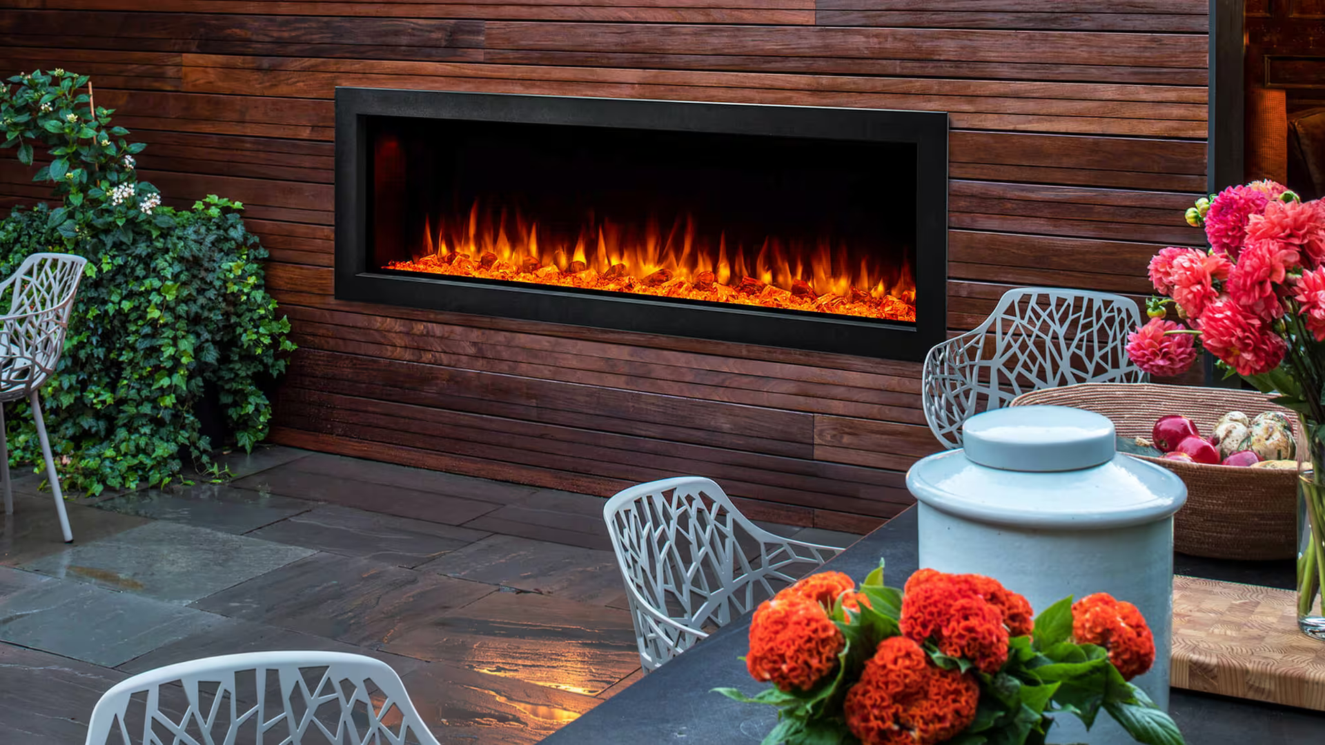 Outdoor fireplace with orange flames, on a wood paneled wall. Outdoor patio with flowers and chairs.