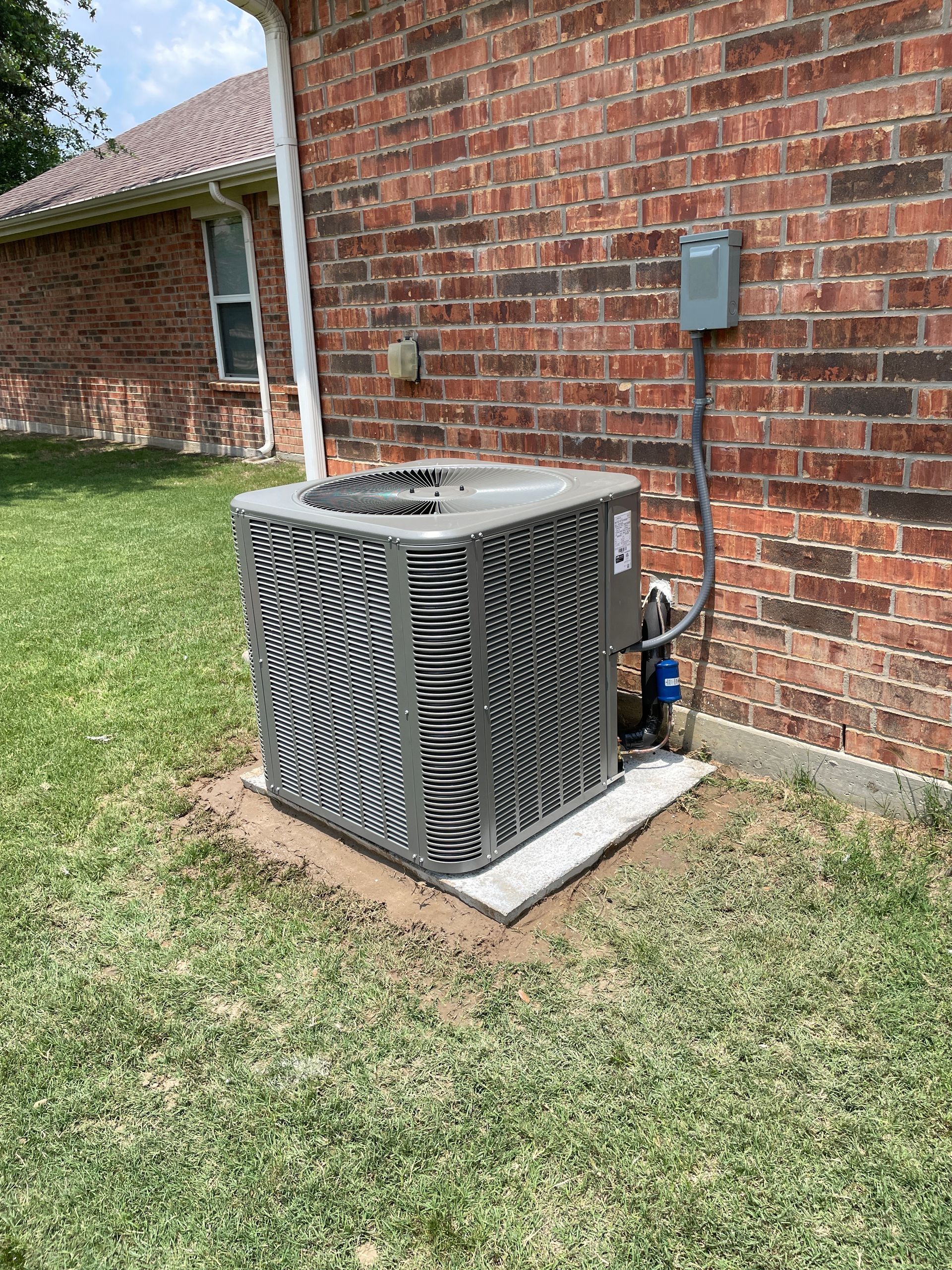 A gray central air conditioning unit sits on a concrete pad against a brick house wall in a grass yard.