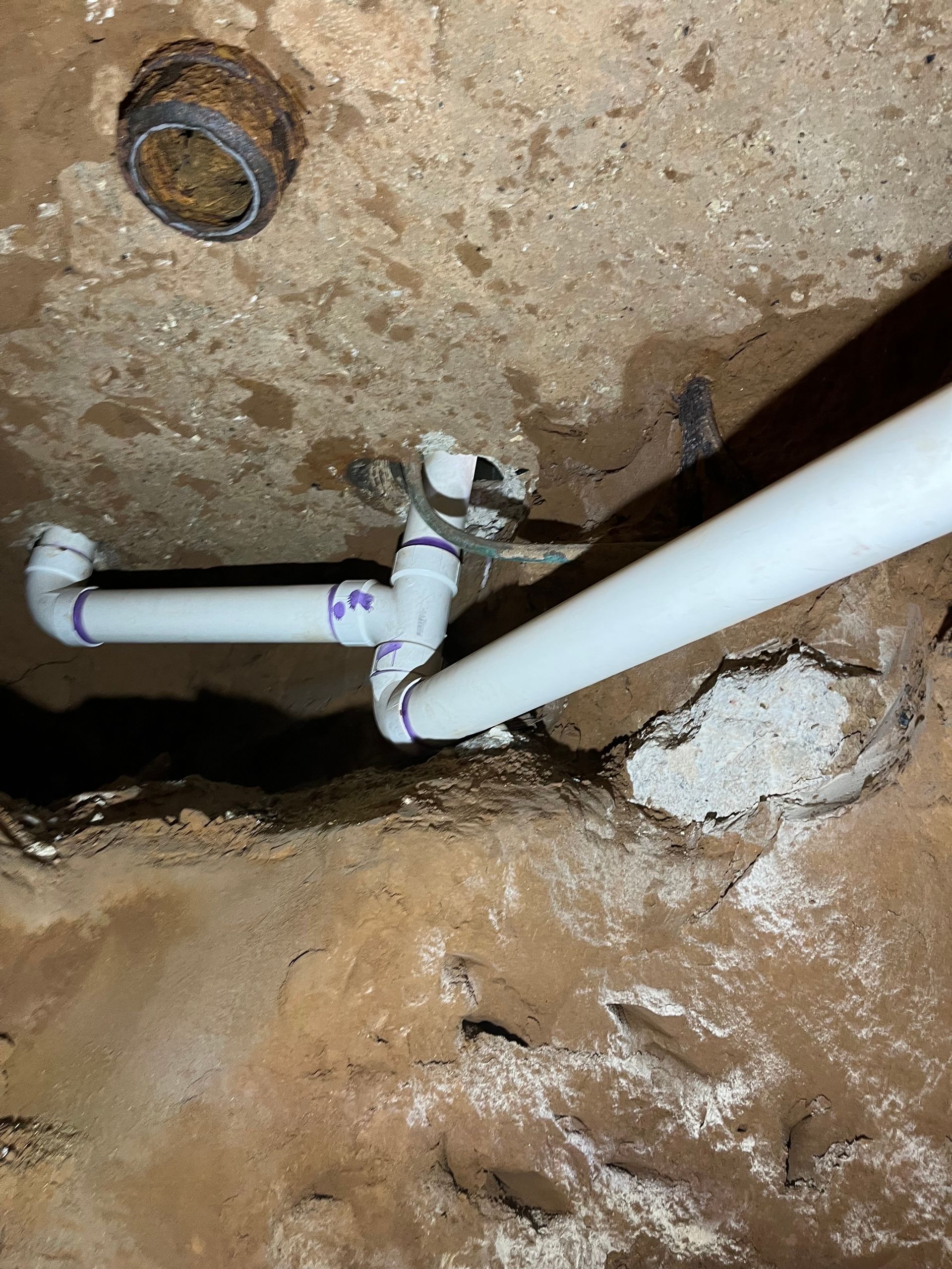 New white PVC plumbing pipes connected to an existing dark drain pipe in an unfinished, dirt-filled basement or crawlspace.