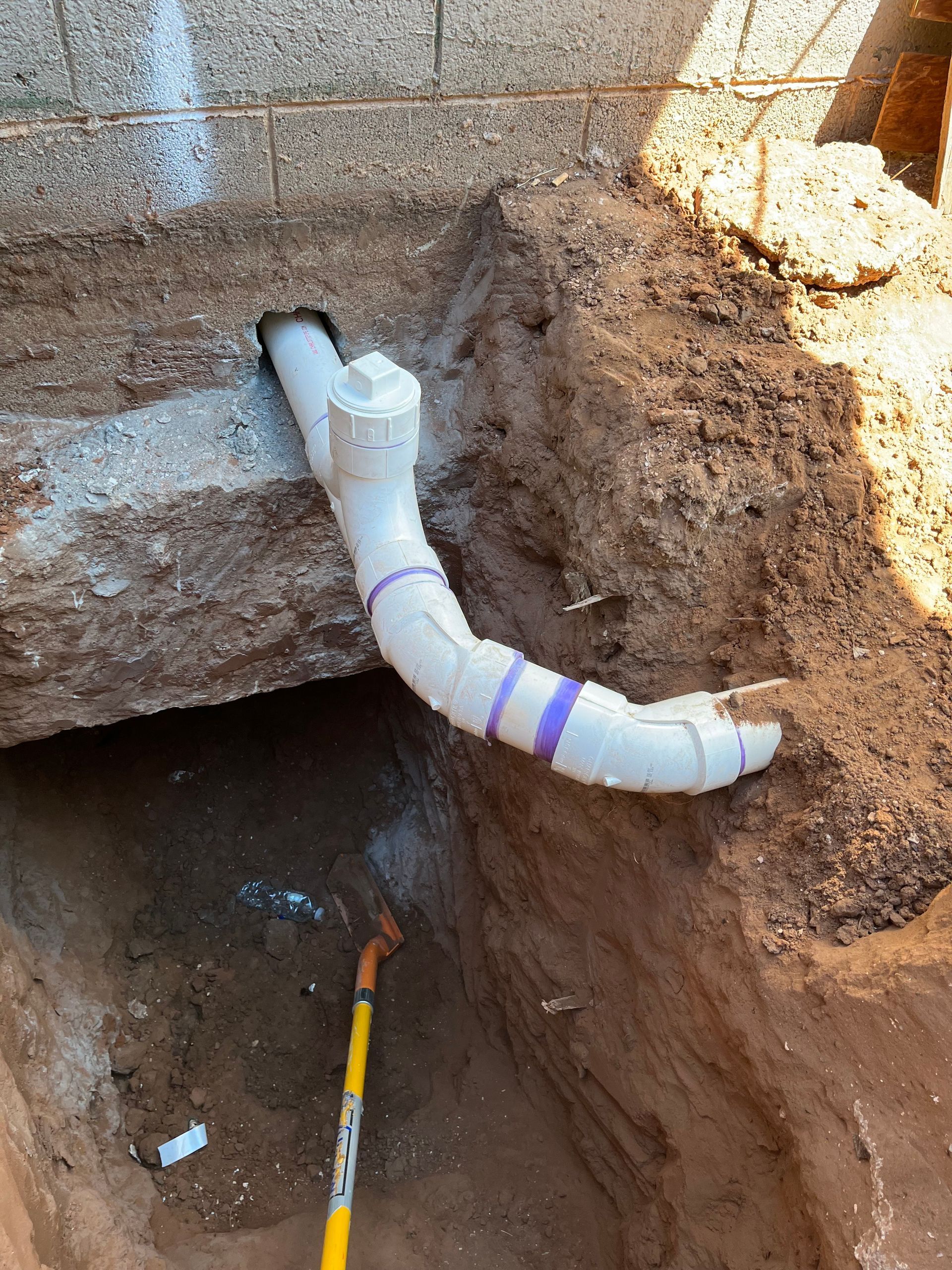 A white PVC cleanout pipe installed in an outdoor trench against a concrete block wall.