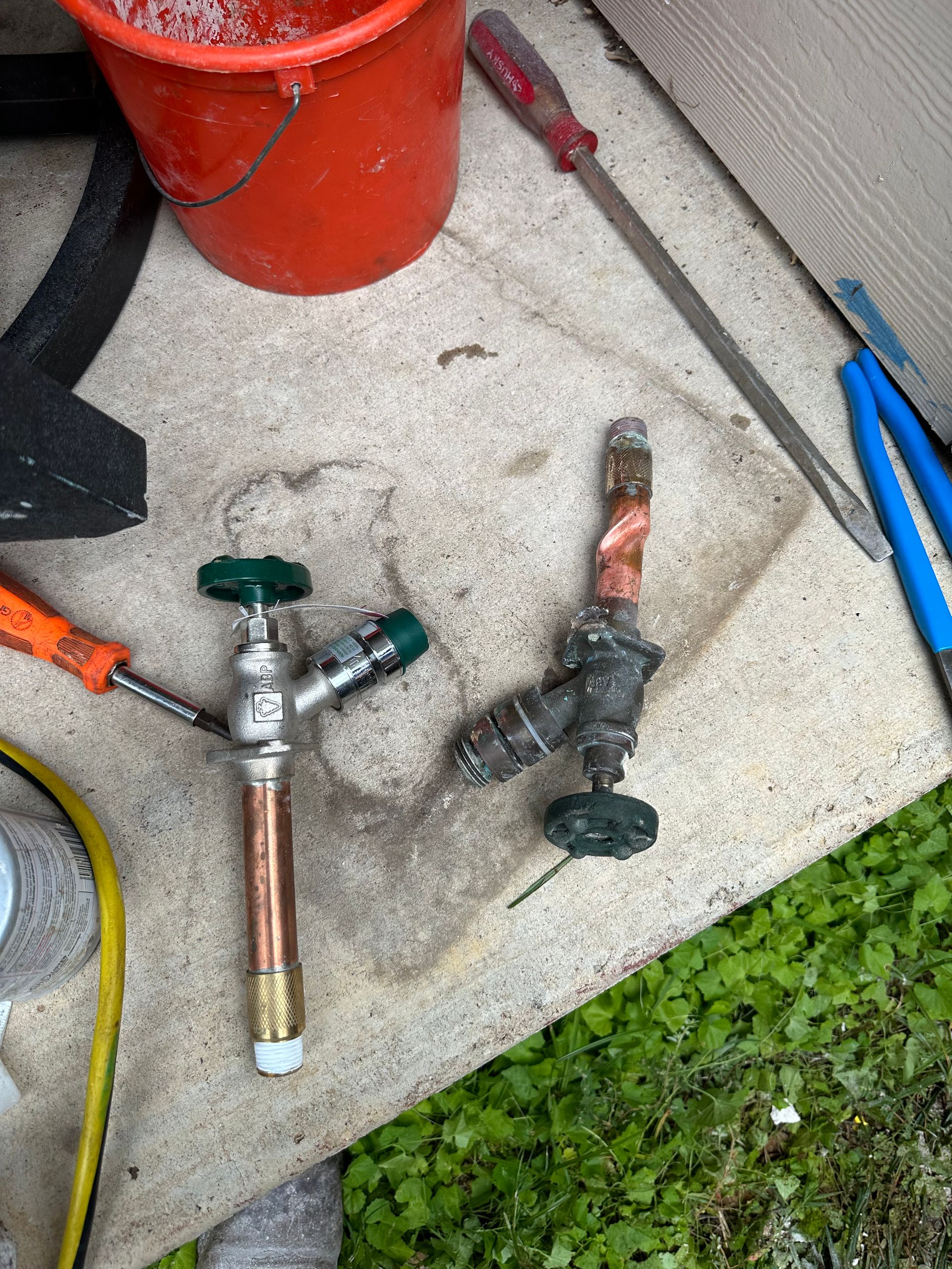 Plumbing work - a new outdoor water spigot and a corroded, removed old one lying on concrete with plumbing tools and a bucket nearby.
