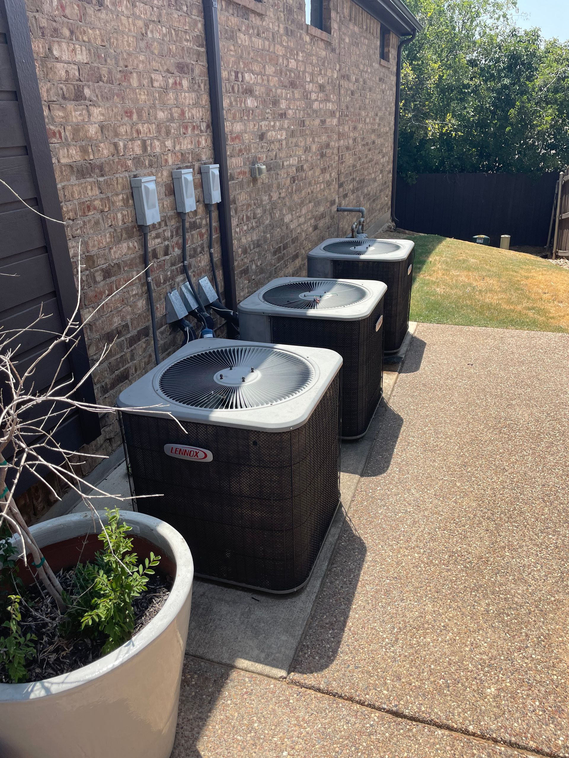 HVAC - Air Conditioning Installation - Three air conditioning units sit in a row along a brick wall on a concrete patio next to a potted plant.