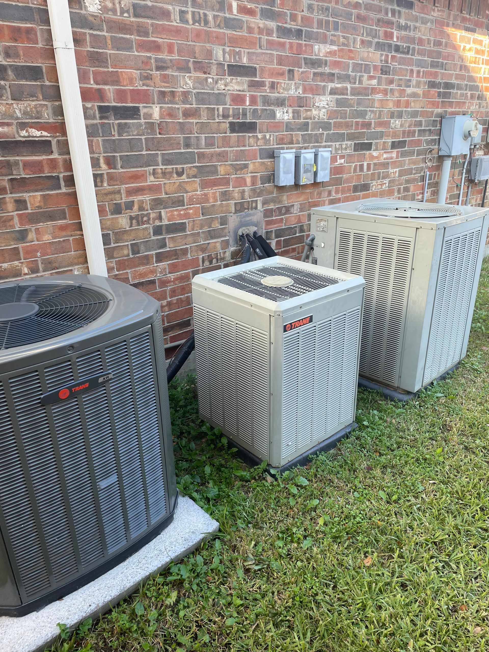 Three outdoor air conditioning condenser units sit on the grass against a brick wall.