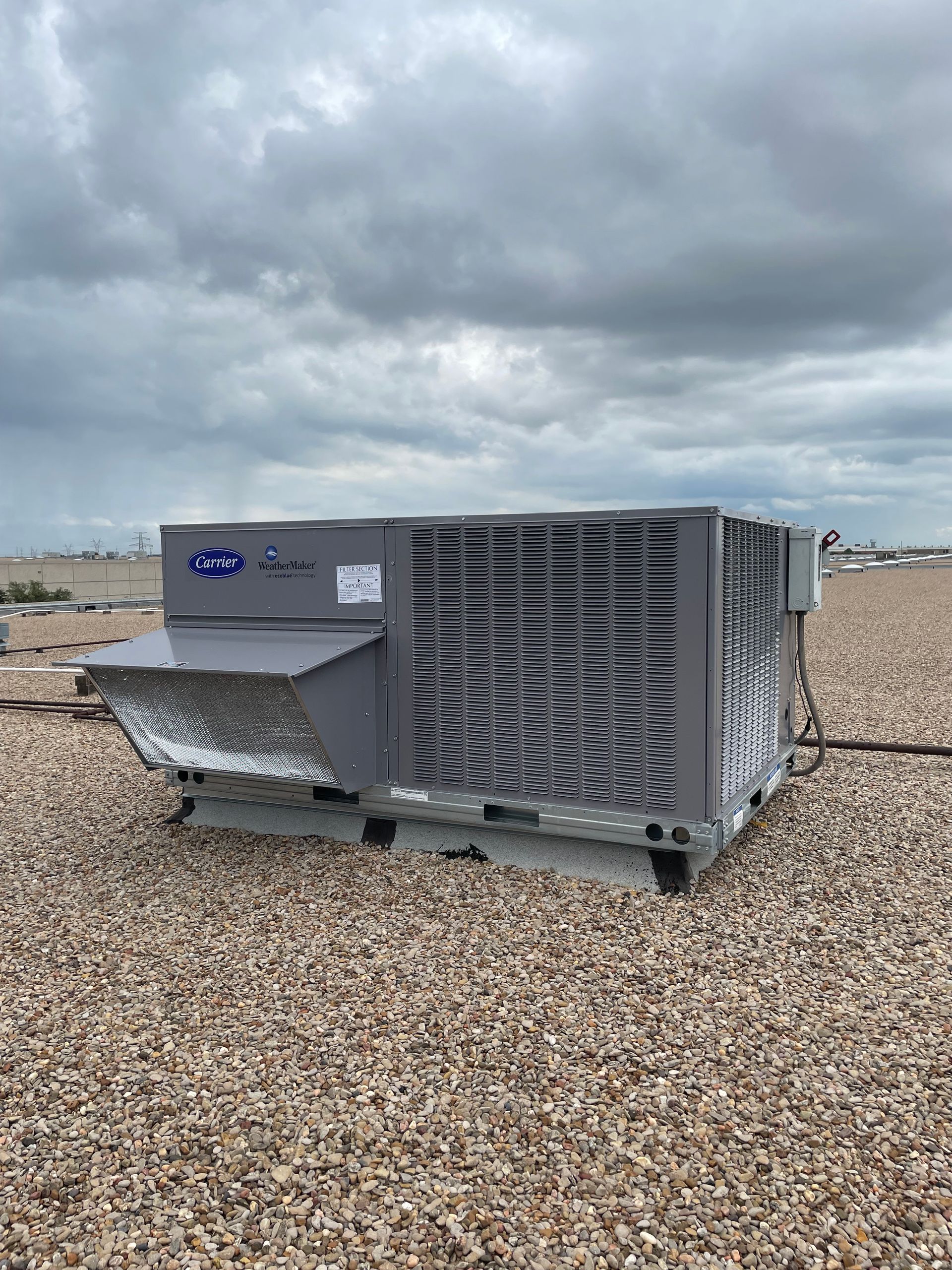 Commercial HVAC job - A grey commercial rooftop HVAC unit sits on a gravel roof under a cloudy sky.