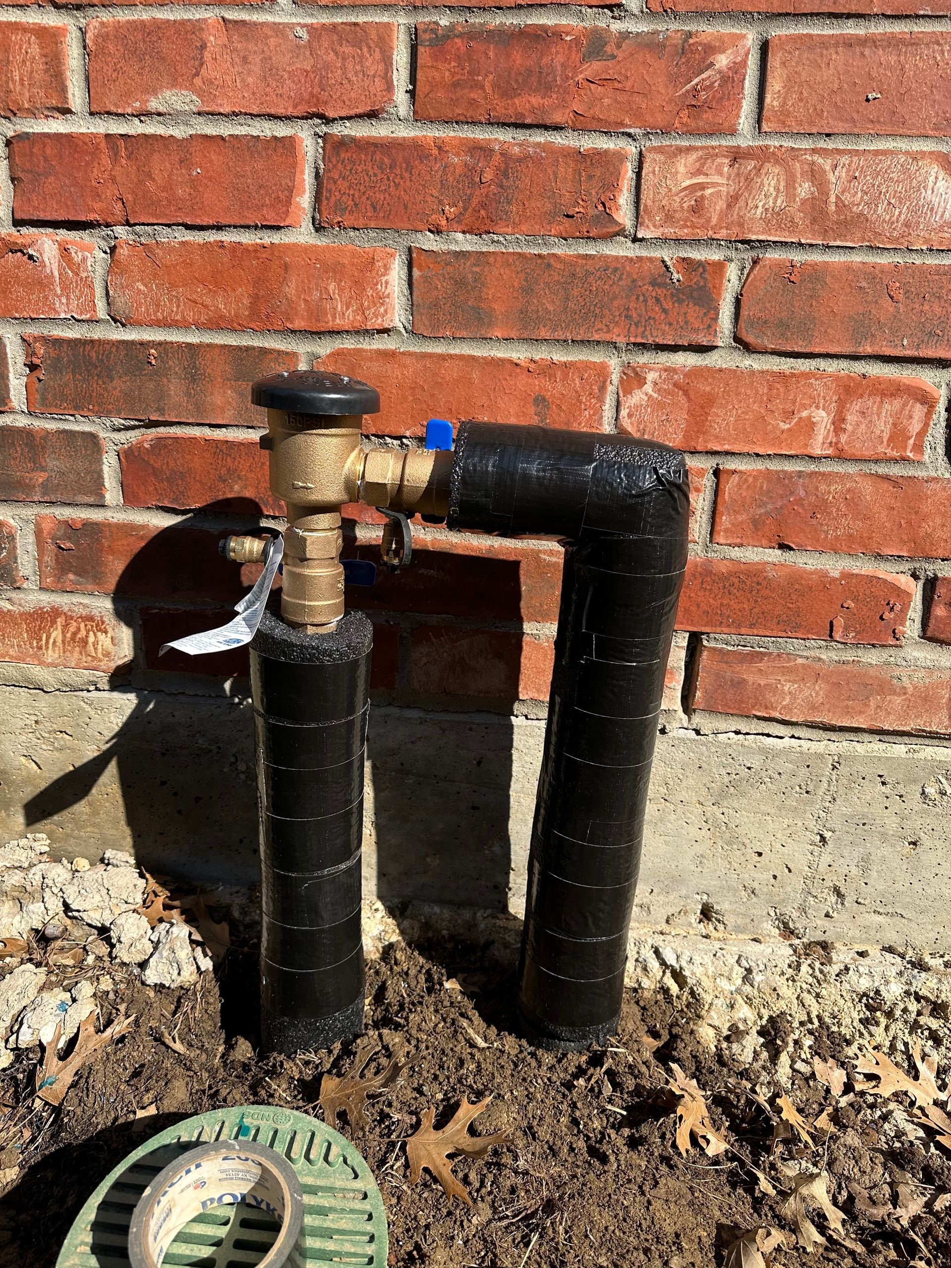 Plumbing work - A brass irrigation backflow preventer assembly insulated with black wrap, installed against a brick wall.