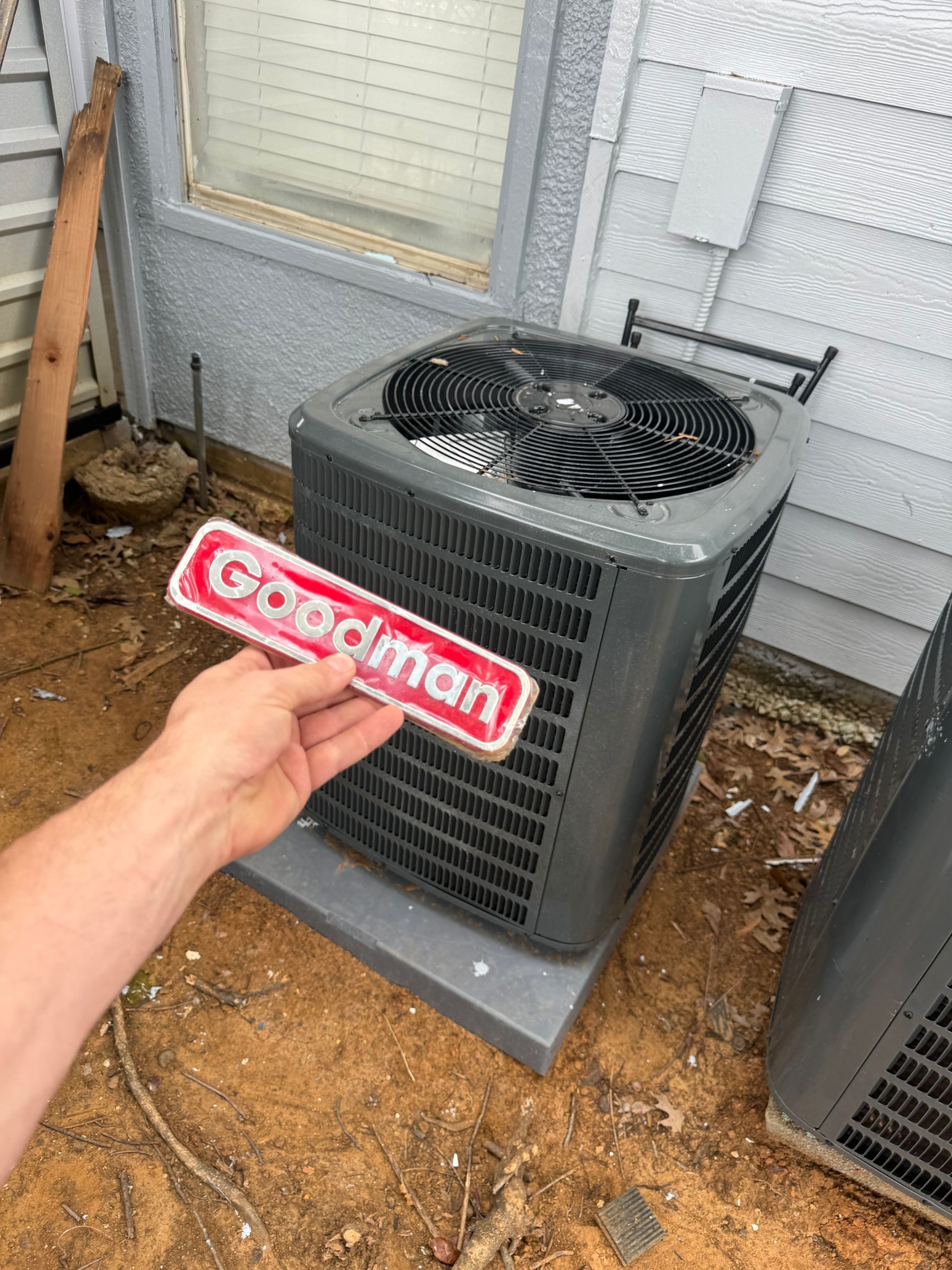 A Serv Tech Pro technician holds a Goodman brand nameplate in front of a gray outdoor air conditioning condenser unit near a house wall.