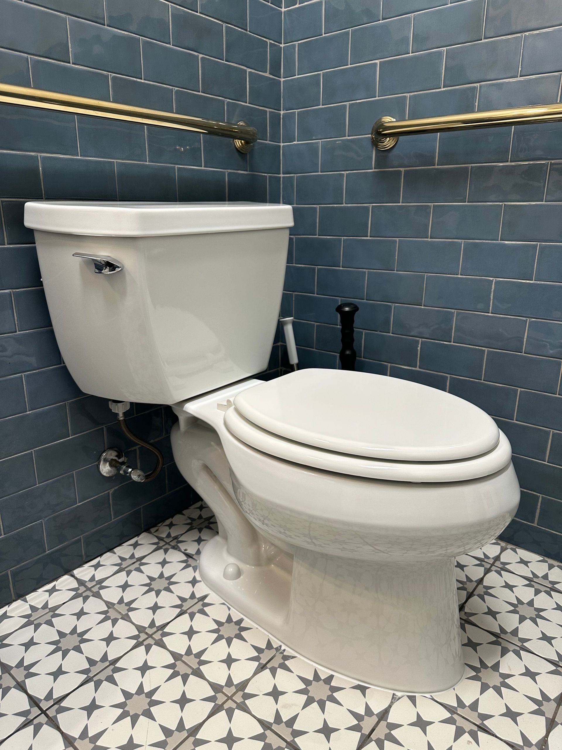 Plumbing job Toilet Installation. A white toilet installed in a corner with blue tiled walls, patterned tile floors, and gold-toned grab bars.
