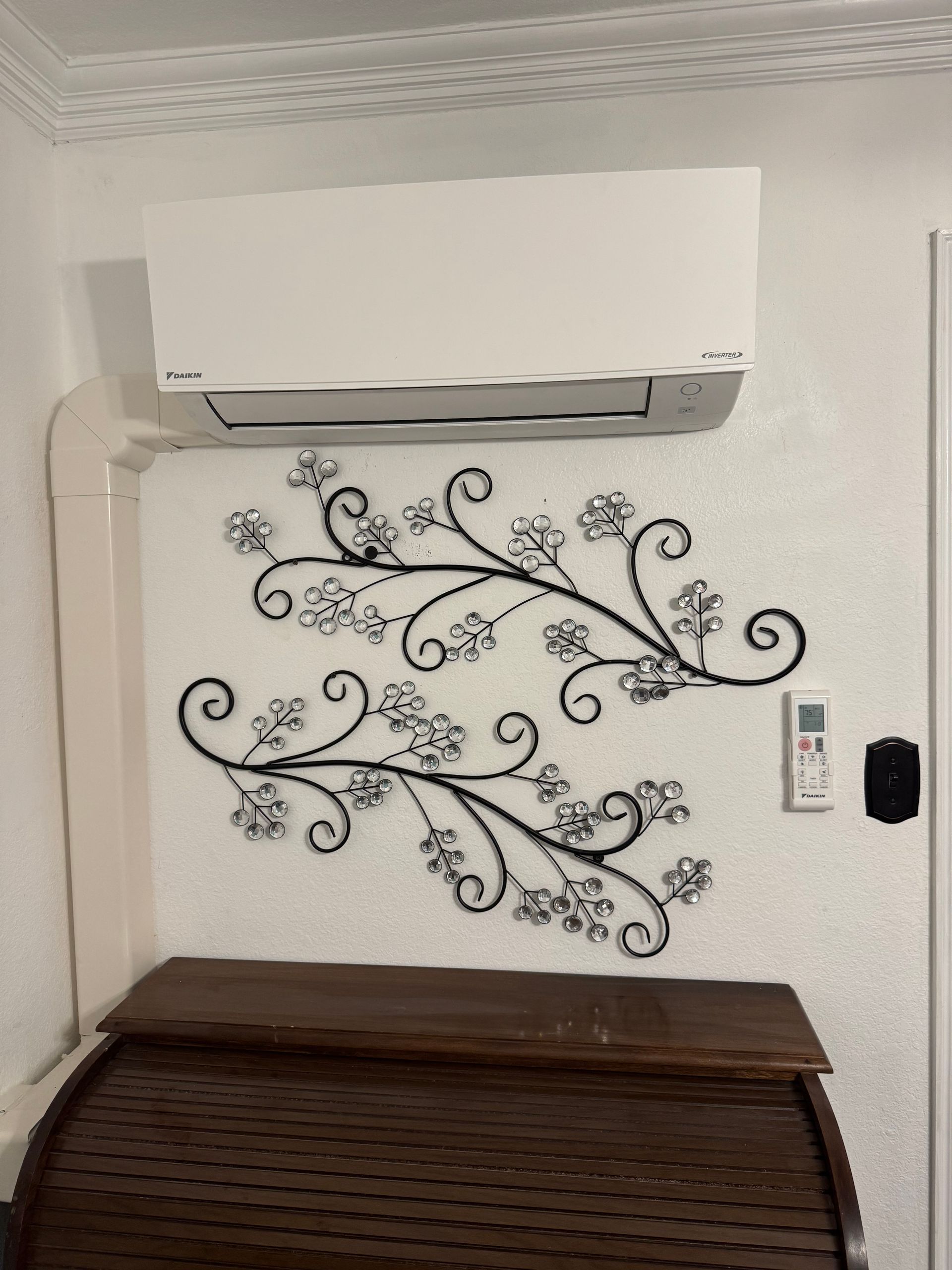 Air Conditioning Installation job. A white wall-mounted air conditioner sits above two black floral metal wall art pieces, with a wooden cabinet below.