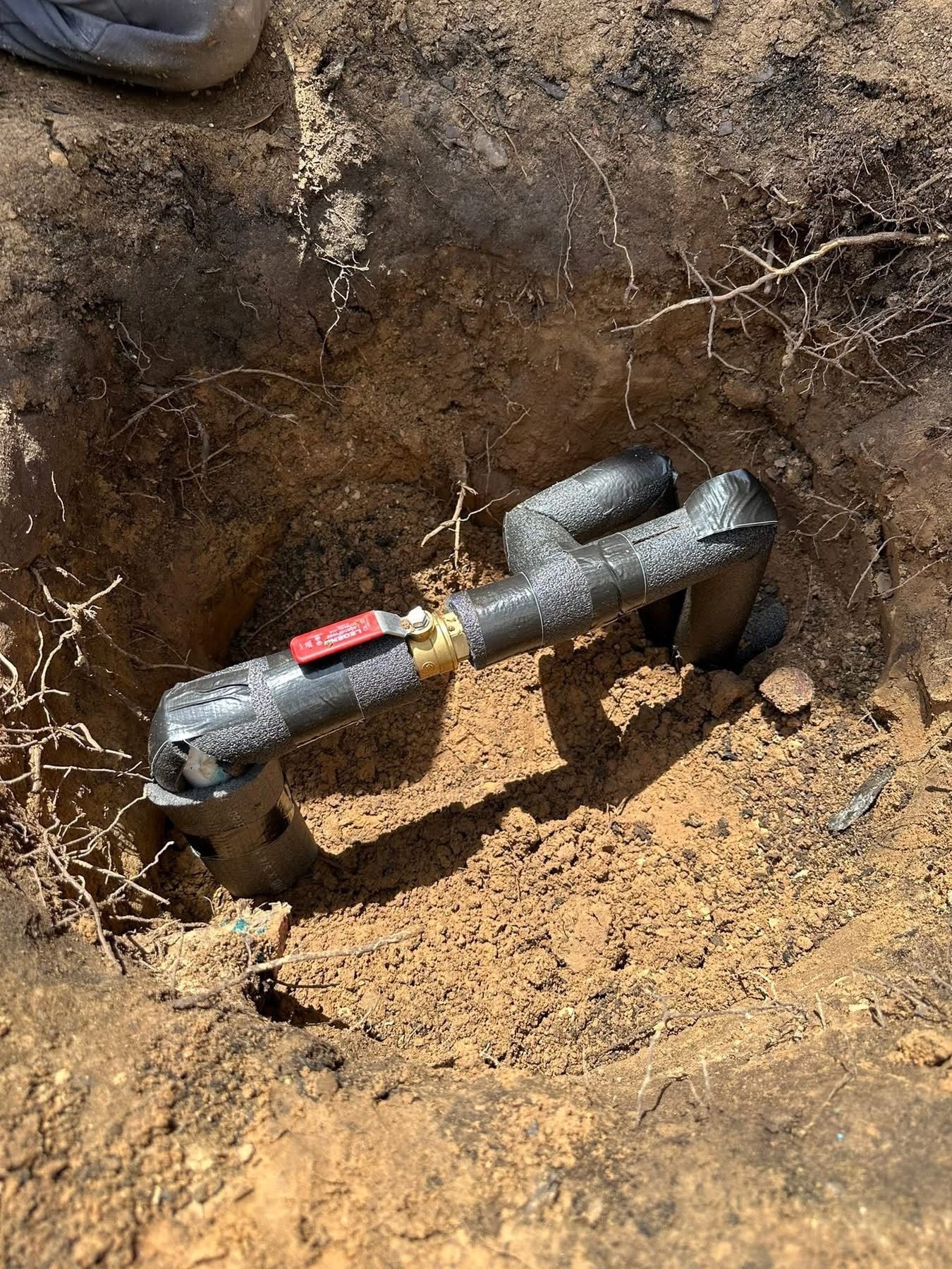 A black metal pipe assembly with a red shut-off valve embedded in a dirt-filled hole in the ground.