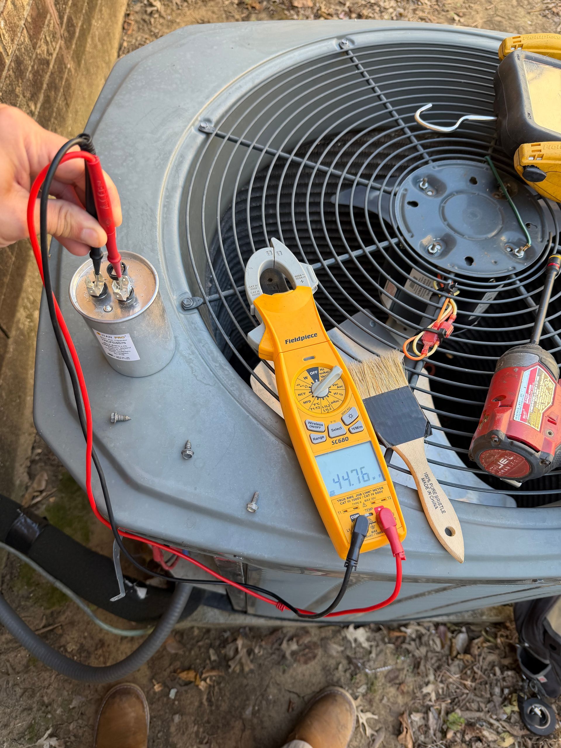 HVAC Service job. A hand tests an HVAC capacitor with a yellow multimeter on top of an outdoor air conditioning unit.
