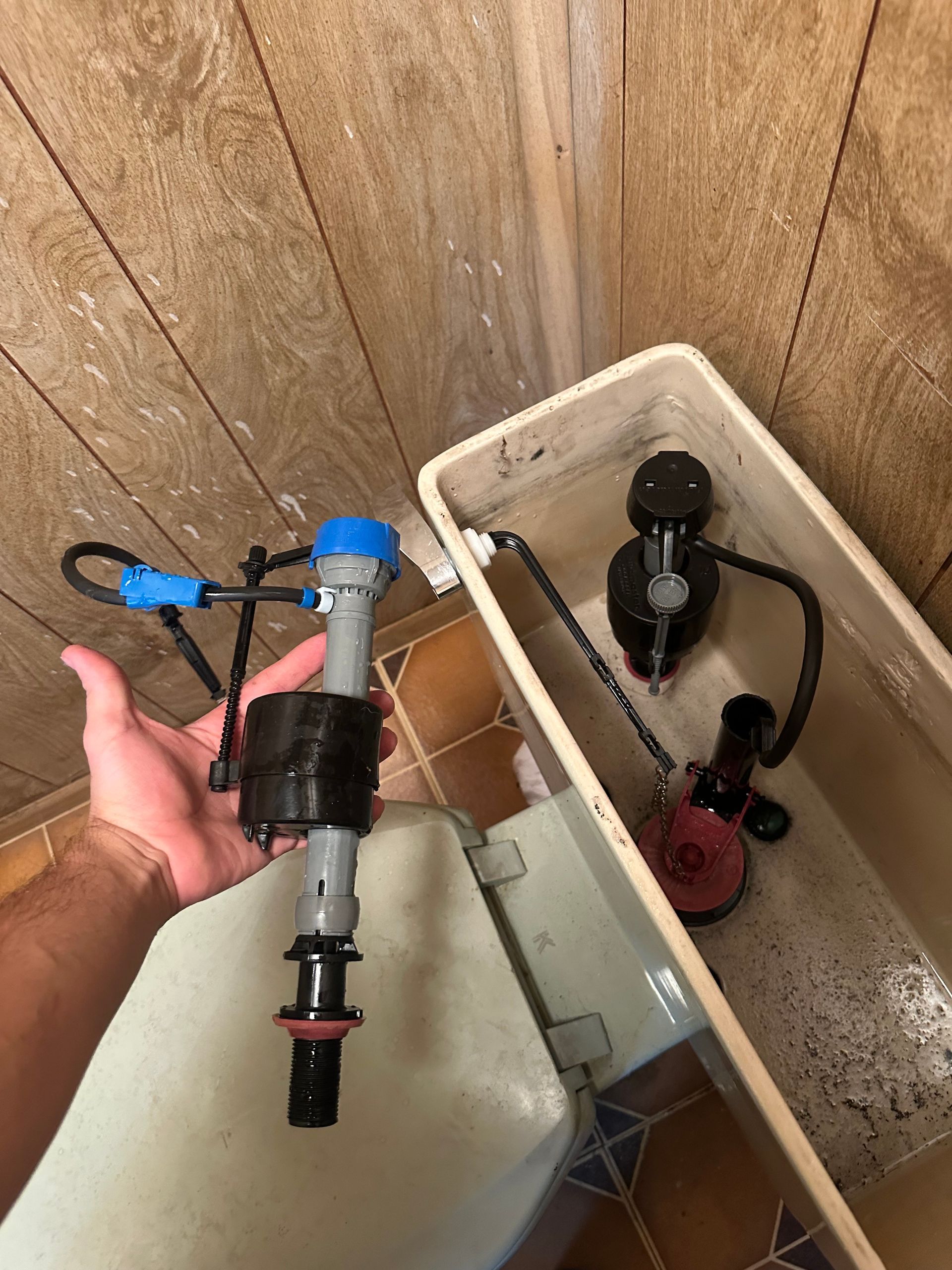 Plumbing repair job. A hand holds a new plastic toilet fill valve beside an open toilet tank containing the old, installed fill valve.