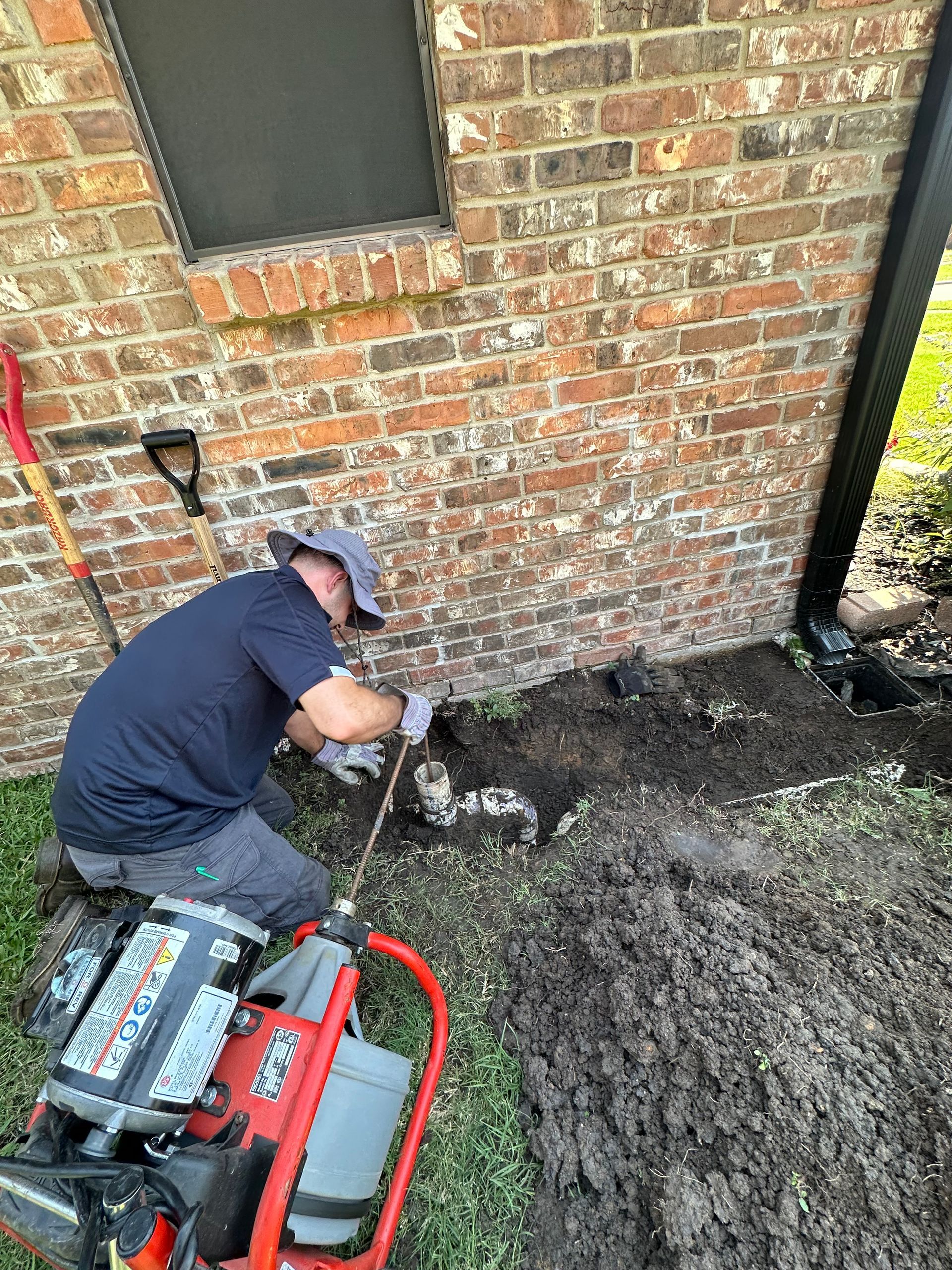 A Serv Tech Pro  worker uses a red power auger to clear a drain pipe in a trench beside a brick house wall.