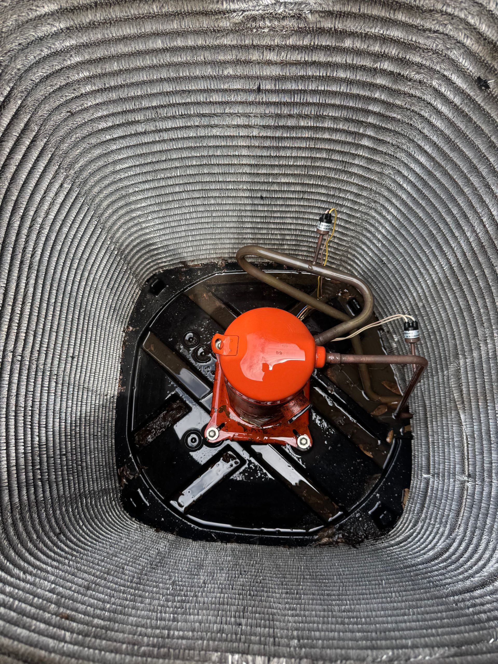 A bright orange, cylindrical compressor sits in the center of an outdoor HVAC unit, surrounded by metal fins.