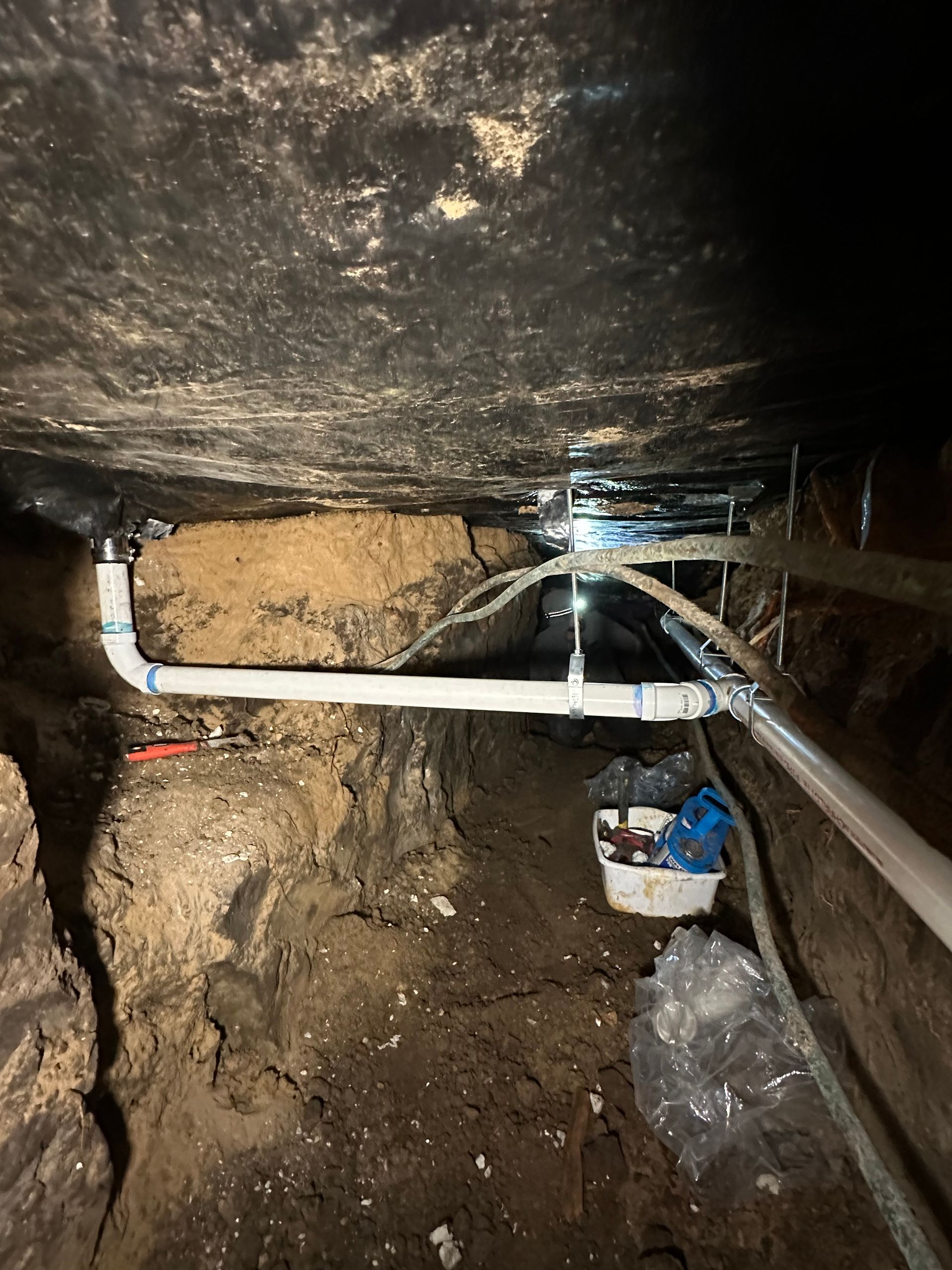 White PVC plumbing pipes run horizontally under a dark crawl space with exposed soil and foundations.