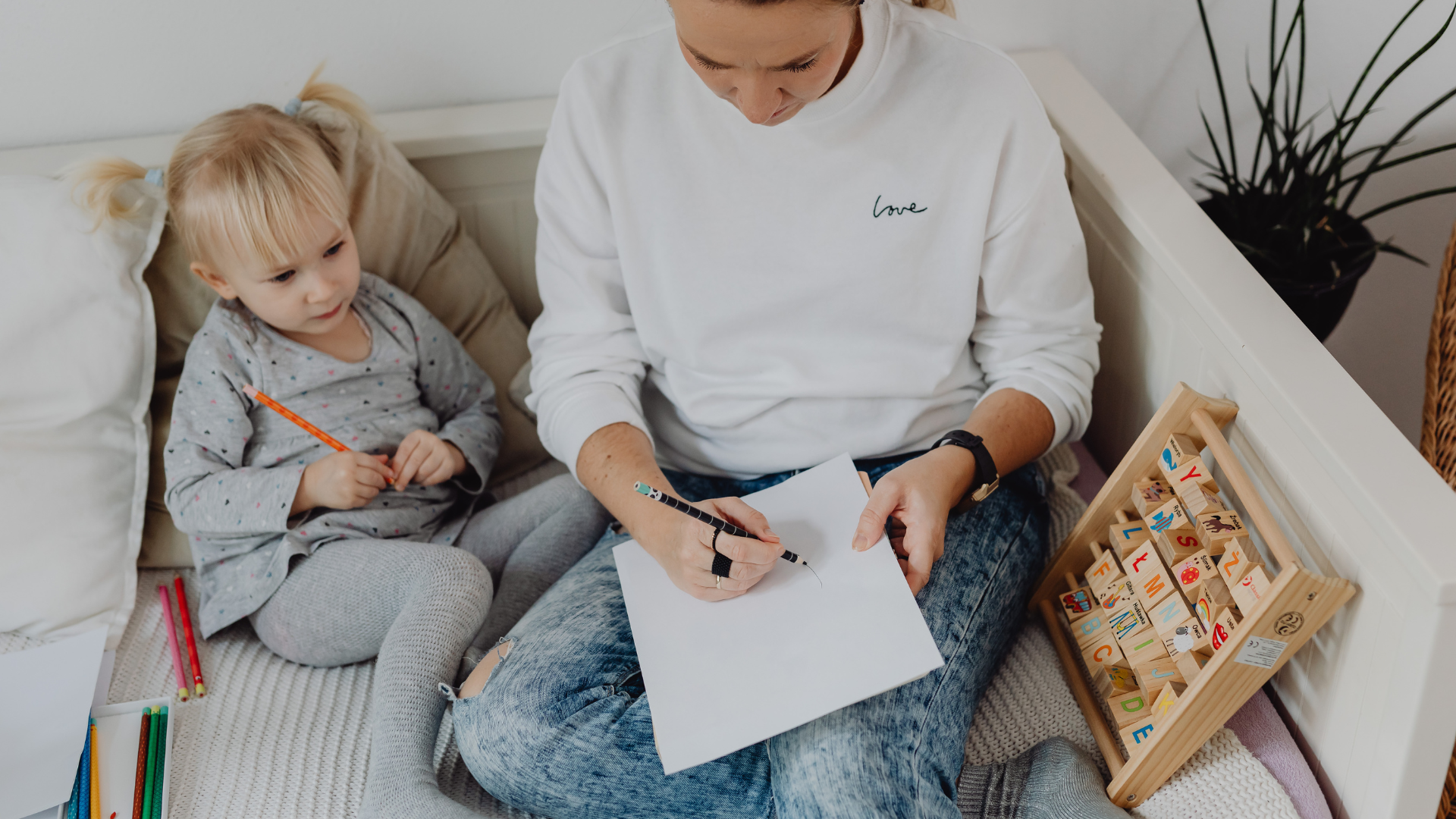 Adult writing on paper beside a child in a cozy room, with a wooden toy on the side.